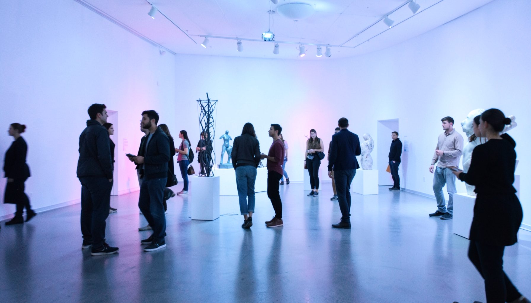 Dimly lit museum gallery with young adults mingling near contemporary sculptures, colored uplighting in purple and blue
