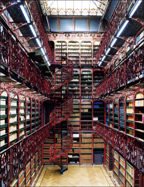 Multi-story library interior with ornate red iron balconies, spiral staircase, and rows of bookshelves lit from a glass ceiling